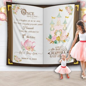 May include: A pink and brown book-shaped backdrop with a castle and teacup illustration. The text reads "Once upon a time... In a kingdom not so far away there lived a beautiful princess named Hannah Today she celebrates her 1st Birthday let's Par-tea MARCH 15, 2022 AND She lived HAPPILY ever after".