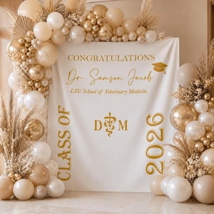 May include: A graduation backdrop featuring a white banner with gold text that reads "CONGRATULATIONS Dr. Samson Jacob LSU School of Veterinary Medicine." The banner is decorated with balloons in shades of white, gold, and beige, and pampas grass.
