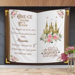 May include: A white book with gold lettering that reads "Once Upon a Time" and a gold castle illustration. The book is decorated with pink and white flowers. The text below reads "In a kingdom not so far away...There lived a beautiful princess named Ariana. Today she celebrates her 1st birthday with her royal guests. October 1st 2023. And she lived happily ever after."