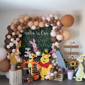 May include: A Winnie the Pooh themed first birthday party decoration set with brown and beige balloons, a green backdrop with the text "Thalia is One", and cardboard cutouts of Winnie the Pooh, Tigger, Piglet, Eeyore, and Roo.