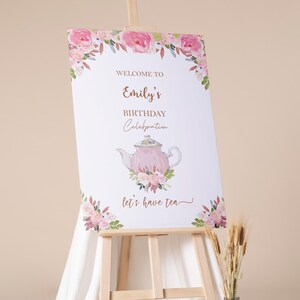 May include: A welcome sign for Emily's birthday celebration. The sign features a pink teapot illustration with floral accents and the phrase "Let's have tea." The sign is displayed on a wooden easel, with a floral border.