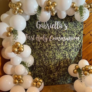 May include: White and gold balloons arranged in an arch with greenery, forming a backdrop for a First Holy Communion celebration. The banner reads "Gabriella's First Holy Communion".