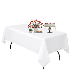 May include: White rectangular tablecloth with a white vase of red tulips, a plate of cookies, and a cake stand with a white cake topped with red berries.