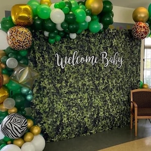 May include: A green and gold balloon arch with a welcome baby sign in front of a green leafy backdrop.