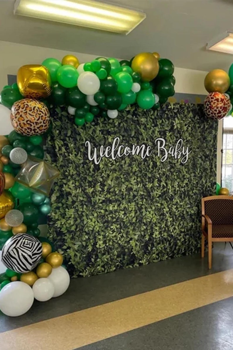 May include: A green and gold balloon arch with a welcome baby sign in front of a green leafy backdrop.