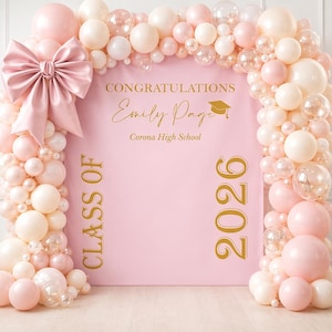 Peut inclure: Un fond rose pour une remise de diplômes avec un grand nœud et une arche de ballons. Le fond porte l'inscription dorée "Félicitations Emily Page Corona High School Classe de 2026". Les ballons sont roses, blancs et transparents.