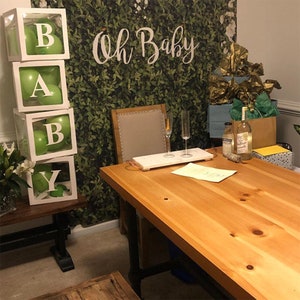 May include: A green and white baby shower decoration with the letters "BABY" spelled out in white boxes with green balloons inside. The boxes are stacked on top of each other. The background is a green leafy wall with the words "Oh Baby" written in white.