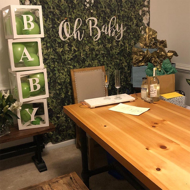 May include: A green and white baby shower decoration with the letters "BABY" spelled out in white boxes with green balloons inside. The boxes are stacked on top of each other. The background is a green leafy wall with the words "Oh Baby" written in white.