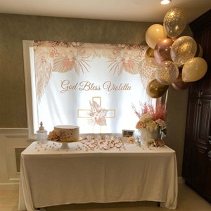 May include: A white table with a white tablecloth is decorated with a white cake with gold accents, a white cross with floral accents, and a banner that reads "God Bless Violetta". The table is decorated with gold and white balloons.