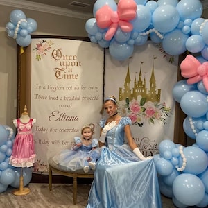 May include: A girl dressed as a princess sits on a bench with a woman dressed as Cinderella. They are in front of a backdrop with a castle and the words "Once upon a time" and "In a kingdom not so far away There lived a beautiful princess named Elliana Today she celebrates birthday with her royal family". The backdrop is decorated with blue and pink balloons.