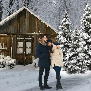 May include: A couple in winter clothing stand in front of a wooden cabin with snow-covered trees in the background.