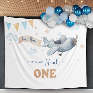May include: A white backdrop with a blue airplane and clouds. The text "Time Flies!" is on a banner attached to the airplane. The text "And Now Noah Is One" is below the airplane. The backdrop is decorated with blue and white balloons.