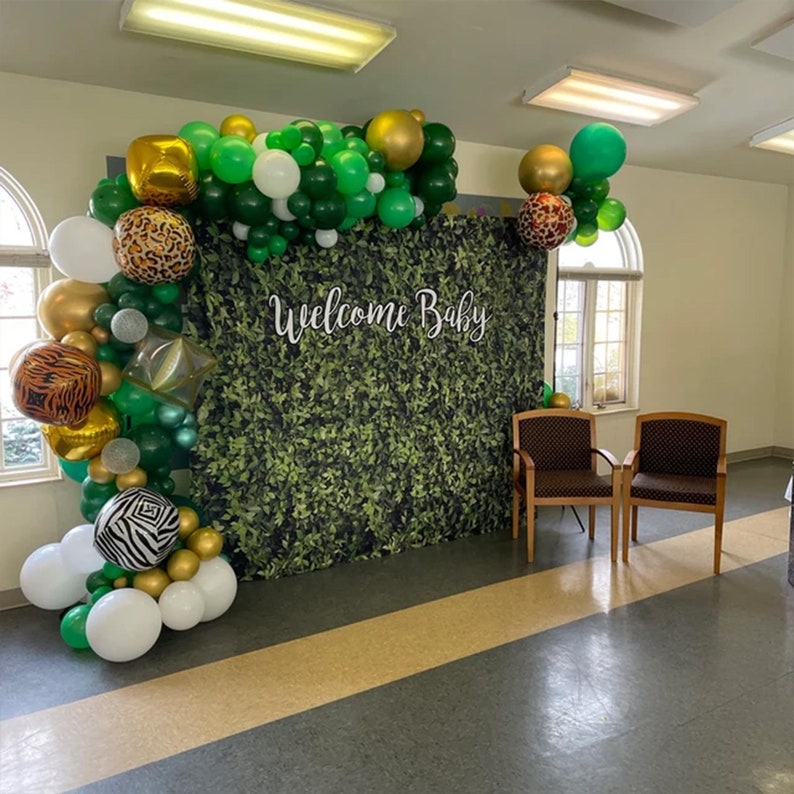 May include: A green and gold balloon arch with animal print balloons and a sign that says "Welcome Baby" in front of a green backdrop.