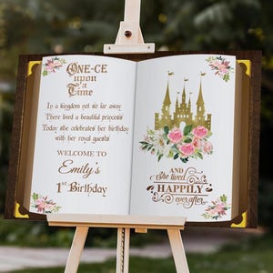 May include: A wooden easel displays a large open book with a fairytale theme. The left page has text that reads "ONE-CE upon a Time" and "Emily's 1st Birthday." The right page features a gold castle illustration and the words "Happily ever after."