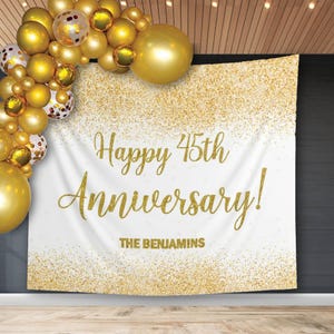 Puede incluir: Una pancarta blanca con detalles de purpurina dorada y las palabras "Happy 45th Anniversary!" en letras doradas. La pancarta también incluye "THE BENJAMINS". Un arco de globos dorados está en la esquina superior izquierda.