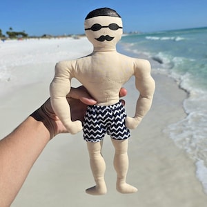 May include: A handmade doll of a swimmer, held in front of a beach scene. The doll has a tan body, black hair, sunglasses, and a mustache. It wears navy and white zigzag patterned swim trunks. The background shows a sandy beach and ocean.