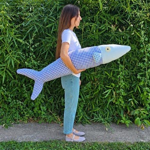 May include: A large, plush fish-shaped pillow featuring a blue and white gingham checkered pattern on the body and tail, with a solid light blue head section, a single visible eye, and a small checkered fin on the side of the elongated body.