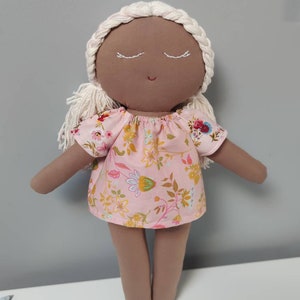 May include: A handmade doll with brown skin, white yarn hair, and closed eyes. The doll is wearing a pink dress with a floral pattern.