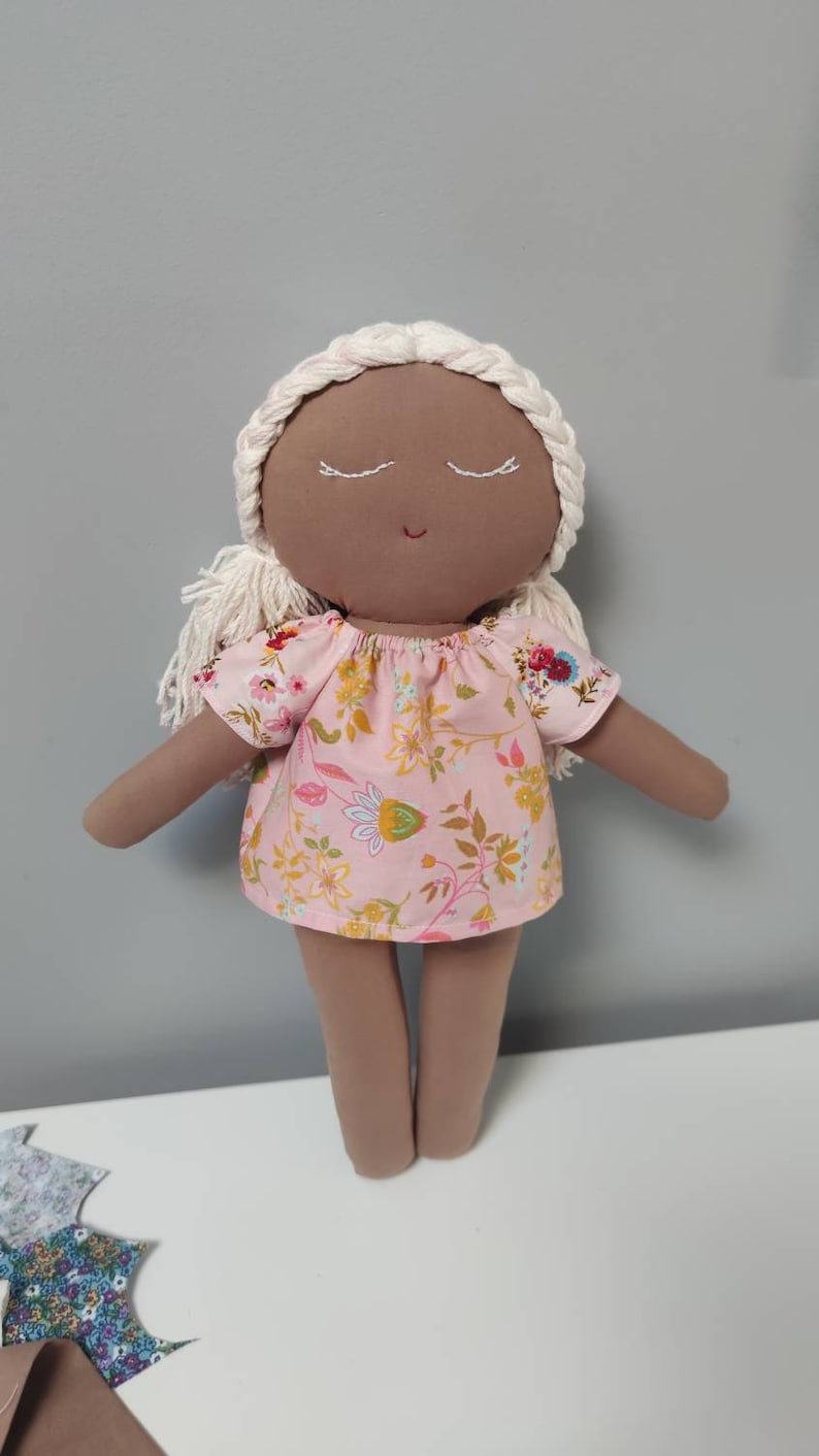 May include: A handmade doll with brown skin, white yarn hair, and closed eyes. The doll is wearing a pink dress with a floral pattern.
