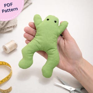 Pode incluir: Um peluche de sapo em tecido verde claro com dois pequenos olhos de contas pretas. O sapo tem uma forma de corpo simples, plana e acolchoada, com braços estendidos e pernas dobradas. A imagem inclui o texto PDF Pattern no canto superior esquerdo.