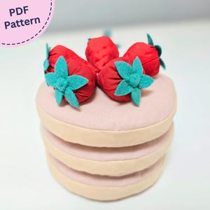 May include: A PDF pattern for a plush stack of three round, light pink fabric pancakes topped with several red fabric strawberries featuring teal green felt leaves. The strawberries are arranged on the top pancake, which has a smooth texture.