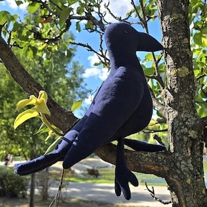 May include: A plush, navy blue crow toy perched on a tree branch. The crow has a large beak and is surrounded by green leaves and a blue sky. The toy is made of soft fabric and has detailed features.