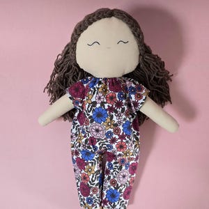 May include: A fabric doll with dark brown yarn hair and a simple embroidered face featuring two curved eyes and a small mouth. The doll wears a one-piece jumpsuit with a colorful floral pattern in shades of blue, pink, red, and yellow on white.