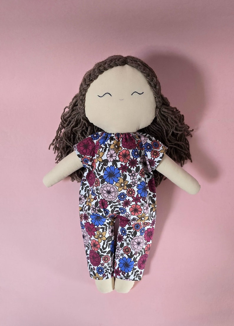 May include: A fabric doll with dark brown yarn hair and a simple embroidered face featuring two curved eyes and a small mouth. The doll wears a one-piece jumpsuit with a colorful floral pattern in shades of blue, pink, red, and yellow on white.