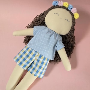May include: A fabric doll with brown yarn hair, a floral crown of blue, pink, and yellow pom-poms, and embroidered facial features. The doll wears a light blue shirt with white polka dots and blue, yellow, and white checkered shorts.