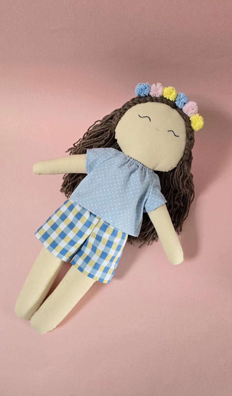 May include: A fabric doll with brown yarn hair, a floral crown of blue, pink, and yellow pom-poms, and embroidered facial features. The doll wears a light blue shirt with white polka dots and blue, yellow, and white checkered shorts.