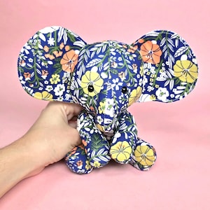 Puede incluir: Un elefante de peluche azul de tela con un estampado floral con flores amarillas, naranjas y blancas. El elefante tiene orejas grandes y una pequeña trompa.