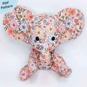May include: A stuffed elephant toy made from fabric featuring a floral pattern with shades of pink, orange, blue, and green. The elephant has large ears, a trunk, and two black button eyes. A circular label in the corner reads PDF Pattern.