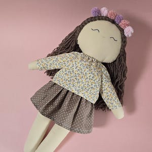 May include: A fabric doll with dark brown yarn hair, featuring a floral print long-sleeve top and a brown skirt with white polka dots. The doll has embroidered facial features and wears a crown of small purple and pink fabric pom-poms.