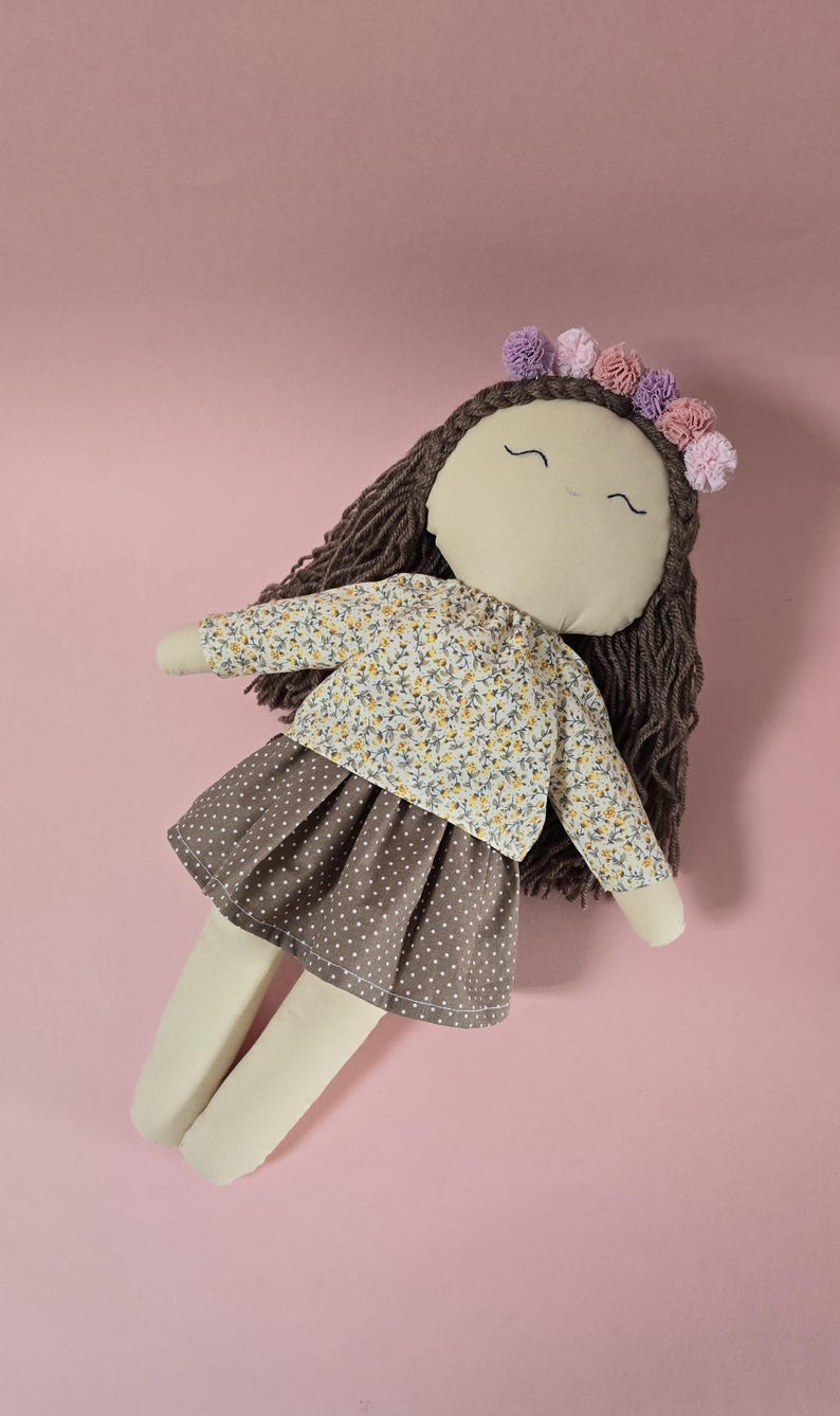 May include: A fabric doll with dark brown yarn hair, featuring a floral print long-sleeve top and a brown skirt with white polka dots. The doll has embroidered facial features and wears a crown of small purple and pink fabric pom-poms.