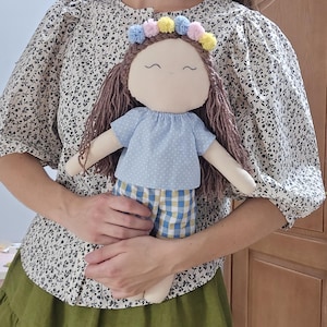 May include: A fabric doll with brown yarn hair, a light blue shirt with white polka dots, and yellow and blue checkered shorts. The doll features a crown of small pastel pom-poms in pink, yellow, and blue, and a simple embroidered face.