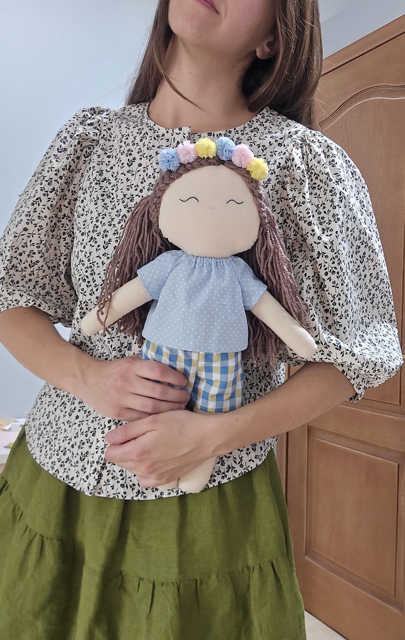 May include: A fabric doll with brown yarn hair, a light blue shirt with white polka dots, and yellow and blue checkered shorts. The doll features a crown of small pastel pom-poms in pink, yellow, and blue, and a simple embroidered face.