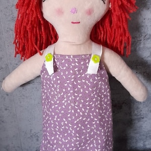 May include: A handmade rag doll with red yarn hair, closed eyes, and a pink nose. The doll is wearing a purple dress with white straps and a floral pattern.