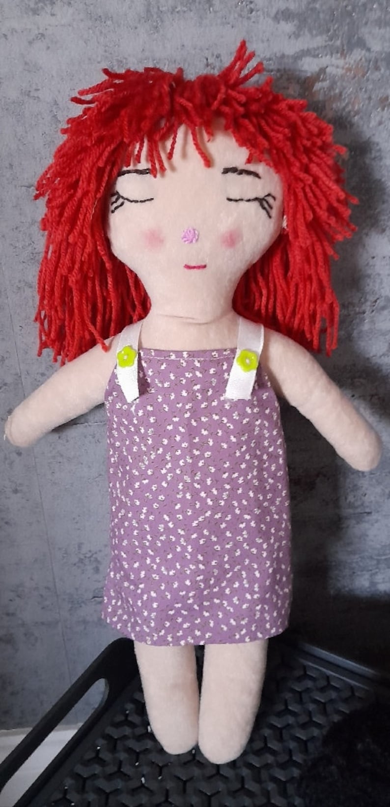 May include: A handmade rag doll with red yarn hair, closed eyes, and a pink nose. The doll is wearing a purple dress with white straps and a floral pattern.