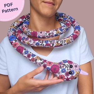 Puede incluir: Un juguete de serpiente de tela relleno con fondo blanco y un patrón floral colorido en tonos rosa, azul y amarillo. La serpiente tiene ojos de cuentas negras y una lengua de color violeta claro. La imagen incluye el texto PDF Pattern.
