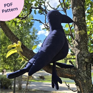 Könnte beinhalten: Ein dunkelblaues Plüsch-Krähenspielzeug mit spitzem Schnabel und weicher Stofftextur. Die Vogelfigur sitzt auf einem Ast. Eine kreisförmige rosa Grafik in der oberen linken Ecke enthält den weißen Text PDF Pattern für eine digitale Anleitung.