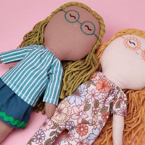 Könnte beinhalten: Zwei handgefertigte Stoffpuppen mit Garnhaaren. Eine Puppe trägt ein grün-weiß gestreiftes Shirt und einen blauen Rock. Die andere Puppe trägt ein Kleid mit Blumenmuster. Beide Puppen tragen Brillen.
