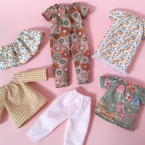 Op de afbeelding: Een set van zes poppenkleding, waaronder een broek met bloemenprint, een geruit shirt, een jurk met bloemenprint, een top met bloemenprint, een rok met bloemenprint en een gestreepte broek.