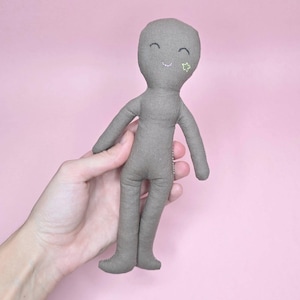 Pode incluir: Uma boneca de tecido cinza com um rosto sorridente e um enfeite em forma de estrela verde. A boneca tem traços simples costurados e uma aparência macia e feita à mão. A boneca é segurada contra um fundo rosa.