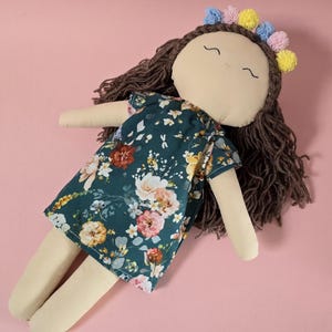 May include: A fabric doll with brown yarn hair, wearing a dark green dress featuring a multi-colored floral pattern. The doll has a beige face with embroidered eyes and a mouth, and is adorned with a headband of small blue, pink, and yellow pom-poms.