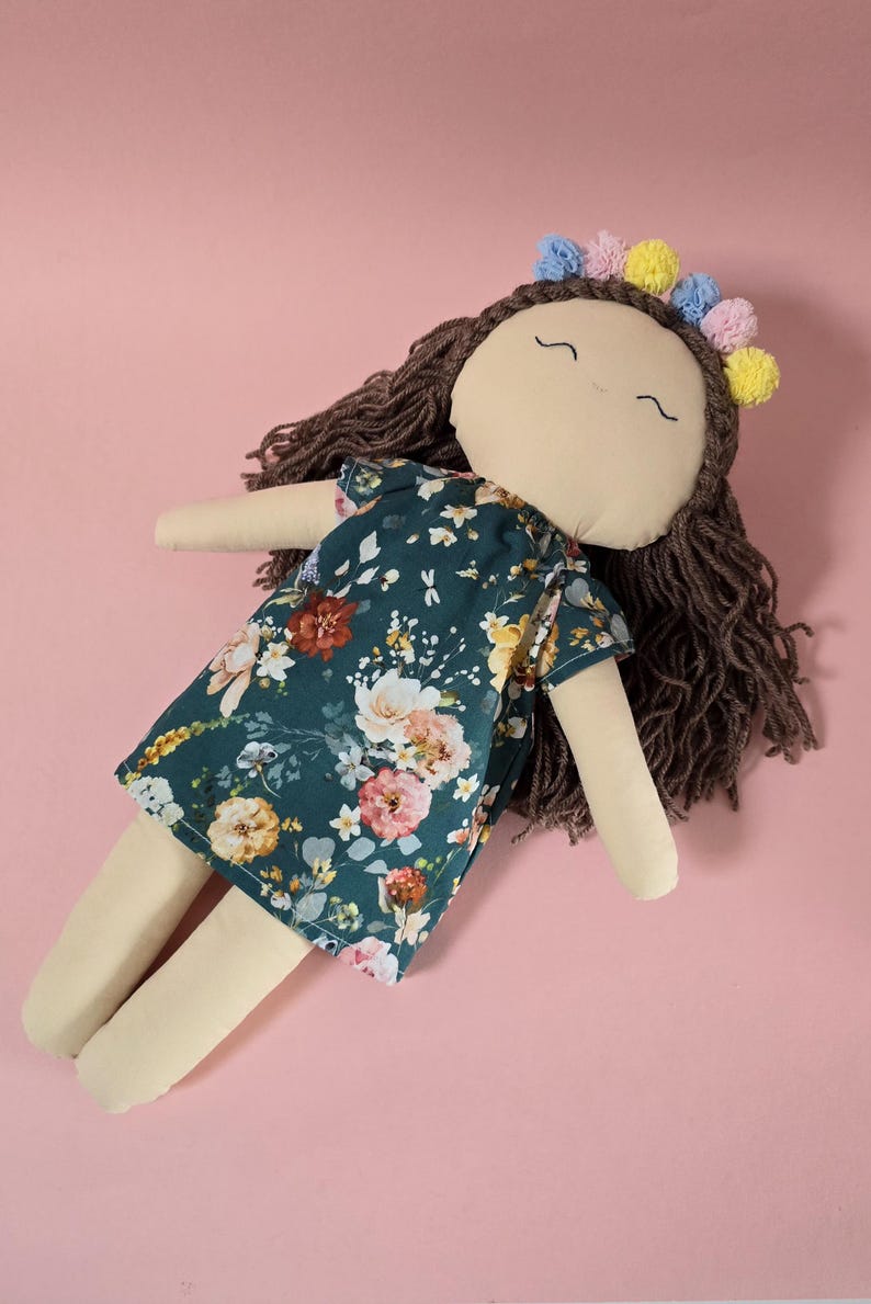 May include: A fabric doll with brown yarn hair, wearing a dark green dress featuring a multi-colored floral pattern. The doll has a beige face with embroidered eyes and a mouth, and is adorned with a headband of small blue, pink, and yellow pom-poms.