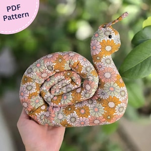 Könnte beinhalten: Ein Stoffschnecken-Spielzeug mit einem wiederkehrenden Blumenmuster in Senfgelb, zartem Rosa und Creme. Die Schnecke hat einen aufgerollten Gehäusekörper, einen aufrechten Hals und einen Fühler mit einem kleinen schwarzen Auge. Text PDF Pattern.