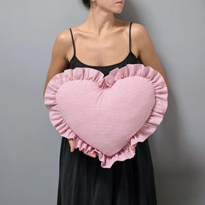 May include: A heart-shaped pillow with a pink and white gingham pattern. The pillow has a ruffled edge and is held up against a gray background.