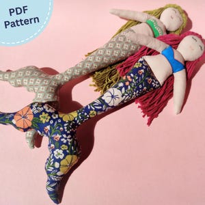 以下が含まれることがあります： 刺繍された顔立ちと毛糸の髪を持つ2体の布製人魚人形。1体はオリーブグリーンの髪と緑のトップが付いたグレーの柄入り尾を持ち、もう1体はマゼンタの髪と青いトップが付いた青い花柄の尾を持っています。PDF Patternというテキストが含まれています。