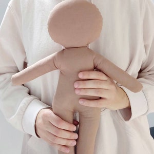 May include: A brown fabric doll with a round head and no facial features. The doll is being held by a person wearing a white hoodie.