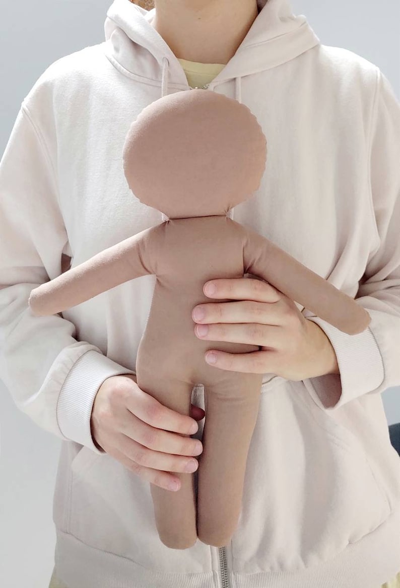 May include: A brown fabric doll with a round head and no facial features. The doll is being held by a person wearing a white hoodie.